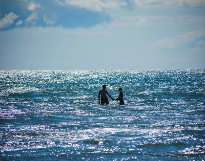 Two hands reaching toward each other over calm water beneath a cloudy sky.